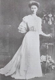 Black and white photograph of Daisy Bates wearing a long white dress. Her hair is tied up and her sleeves are layered. She is looking at the camera while standing with an arm behind her back.