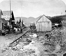 A black and white photo of a Barkerville street. On the left, a series of single-story houses follow the road into the distance; the closest one bears a sign for the "Barnard Express." In the centre, the road edges onto a shallow ditch, flanked by rocks and mining rubble. A lone one-story building with a sloped roof stands at the end of the ditch, with its windowless side wall facing the viewer. To the right of the photo, more single-story structures are seen some 50 metres away.