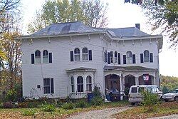 A white house with black shutters on its rounded windows. There is a white van parked in a driveway to its right, and a banner with "ny" in white on a red-and-blue background