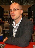 A suited bald man looks directly into camera at a signing event.