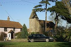 Stone church with square tower in the background partially obscured by trees. To the left is a pink painted house with red roof and in the foreground a car and grass area.