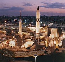 Against a twilight cityscape stand two rockets, a space capsule, a rocket engine, and a lunar lander. They are illuminated with floodlights.