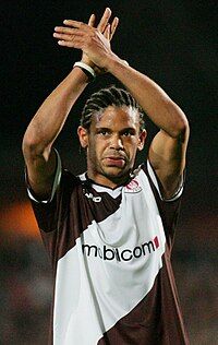 African man in a diagonally striped brown-and-white jersey with a sponsor logo across the chest. His hair is cornrowed, and his arms are crossed above his head.