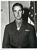 Head and torso of a young white man with neatly combed hair wearing a dark military jacket with a wide belt and a few ribbon bars on the left breast. A star-shaped medal hangs from a ribbon around his neck.