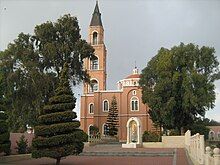 Orange coloured church with steeple visible between trees and a courtyard in the foreground