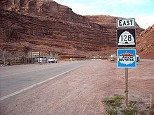 Signpost assembly against a red sandstone backdrop. The signs on the post read, "East, 128 (in a beehive shaped shield), and Scenic Byway".