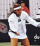 A woman in all white dress, white jacket, and white headband, which she is holding a blue tennis racket