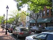 A sunny downtown setting with several cars parked along both sides of a street, with a row of storefronts on one side and a red bricked sidewalk with tall lamposts on the other.