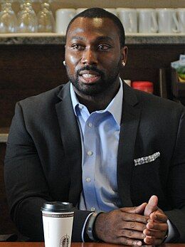 Kerry Carter sits at at table. He's dressed in a dark sport coat and is wearing a blue oxford shirt, unbuttoned at the collar.