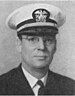 Head of a middle-aged white man wearing a white peaked cap, wire frame glasses, and a dark jacket over a shirt and tie.