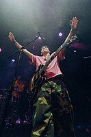 An image of Clinton Kane from a below point-of-view. He is wearing green camo cargo pants and a reddish pink short-sleeve button-up shirt. He has a guitar hung on his shoulder, and is raising his hands out forward and above his head.