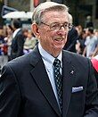 A man wearing a dark blue sports coat with a ribbon-shaped pin on his lapel