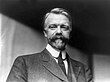 A black-and-white of a man with a mustache wearing a suit and a tie