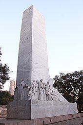 The rectangular base of a cenotaph. An angel is carved on one end. On the side are carvings of several men, shown wearing bucksin or 19th-century suits. Many hold guns or knives; at the far end, one operates a cannon.