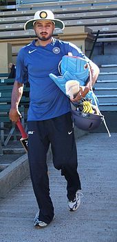 Man with black beard in a turban, wearing a white broad brimmed sunhat, is wearing navy tracksuit pants, blue T-shirt, and carrying light blue batting pads, a cricket bat and a helmet. Spectator seats can be seen in the background.