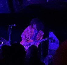 Moon Walker performing on stage. Harry Springer is playing the electric guitar and wearing a checkered suit.