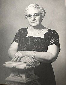 Formal waist-up black-and-white photograph of a white woman in a black lace dress, wearing pearls, her white hair curled, wearing cat-eye glasses. She is leaning on a faux marble Grecian-style column.