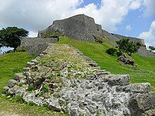 A photo of a large grey castle on a grassy hill dotted by stone ruins and small trees.