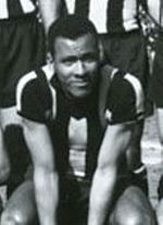 A young man sitting down, wearing a striped shirt. Behind him, three men wearing the same shirt and dark shorts are partially visible