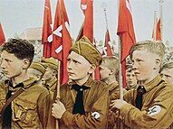 Young boys, wearing brown Nazi uniforms, on parade with red swastika banners