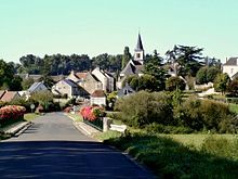 A general view of a village in tiers on the side of a wooded hill with a river in the foreground.