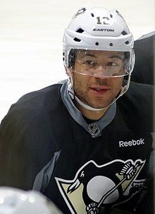 Upper body of a man looking to his right. He is in a black jersey with a stylized penguin logo on his chest and is wearing a white helmet with a visor.