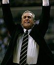 A man raising his hands in the air during a basketball game. He is wearing an open jacket with a white shirt and striped tie.