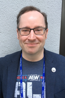 A smiling white man with dark hair, wearing glasses, a dark jacket, a dark t-shirt, and a lanyard