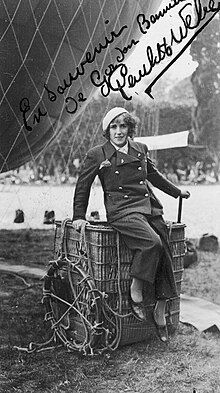 Young woman sitting on the edge of a hot air balloon basket that is set on the ground. She's wearing pants, a double-breasted jacket and a beret. There is a part of a hot air balloon looming behind her.