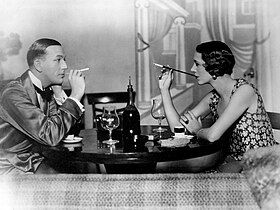 young white man, clean shaven, facing a young white woman with bob cut hair across a small round table set with wine glasses and demitasse cups; both are smoking using cigarette holders