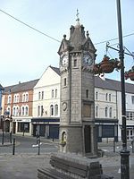 A tower-like structure in stone with a clock face on each side. Above each clock is a gable and at the summit of the structure is a spire.