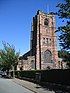 The west end of a Gothic Revival style church built in different coloured sandstone, showing a tall tower with a flagpole; in front is a hedge and to the sides are trees