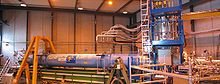 A warehouse interior containing a long blue cylinder surrounded by scaffolding and plumbing.