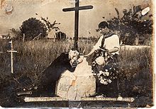 Black and white photo showing a man kneeling on a grave decorated with a cross, with another man standing next to him, holding a wreath of flowers.