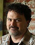 A portrait of a man with curly black hair and a salt-and-pepper beard facing the left of the screen, eyes turned towards the viewer