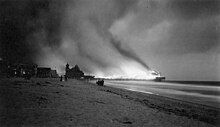 Burning pier and billowing smoke in black and white photograph taken around dusk