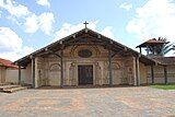 A church and bell-tower in frontal view. The whitish facade is decorated with motifs painted in orange. A wooden cross is positioned at the top of the roof.