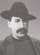 A black and white photograph of Jack Bates. He is wearing a black trilby and is sporting a thick, dark mustache.