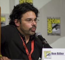 A man wearing a black/brown shirt speaks into a microphone while looking straight forward.