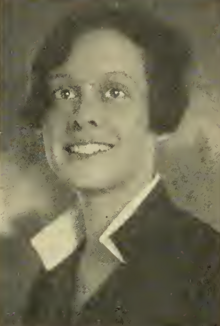 A smiling young woman with tan skin and dark bobbed hair; she is wearing a dark blouse with a white-lined standing collar