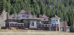 An elaborate stone and wood building with many peaked roofs and chimneys with a wooded hill behind it.