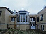 An oriel window topped by a cross protrudes from a building clad in beige brick.