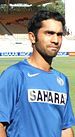 Brown-skinned young man, not clean shaven, wears a sky blue shirt with the words "SAHARA" on it stares forward. In the background is lawn of a sporting ground and a grandstand.