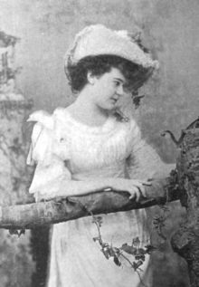 A young white woman posted with her arm on a tree branch. She is wearing a white dress and hat; her dark curly hair is arranged in an updo.