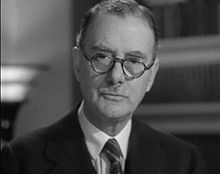 A black and white head and shoulders photograph of a middle-aged man looking at the viewer. He has with round glasses and a moustache and is wearing a dark suit jacket, white shirt and stripped tie.