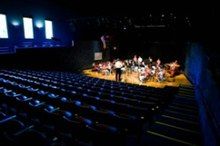 Empty blue seats and a small orchestra on a lighted stage, as seen from the rear of an auditorium