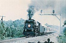 A black steam locomotive pulling a long rake of passenger coaches, with many trees and a signal tower in the background