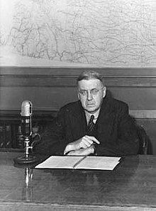 An unsmiling man with round-rimmed glasses and greying hair sitting before a microphone with a cerlox-bound book open in front of him