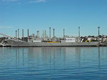 A small warship tied up alongside at a wharf. Her communications and radar masts have been crudely downsized, she carries no weapons, and several large squares have been cut into the ship's hull.