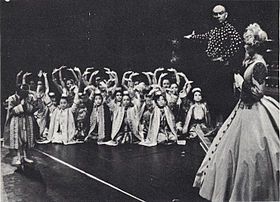 Black and white photo of a theatrical scene: a middle-aged man with a shaved head and imposing presence has his right arm extended to introduce a group of children in Asian dress to a woman in a crinoline dress and bonnet in the foreground at right, who is partially turned upstage. The children are mostly kneeling and have their arms raised in greeting; one child (probably Crown Prince Chulalongkorn) stands and bows.